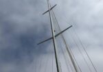 Upward view of a sailboat mast with spreaders and rigging against a cloudy sky.