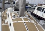 Deck of a sailboat in a marina with beige non-slip panels, furled mainsail cover, winches and lines visible on the cockpit deck.