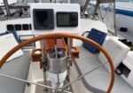 Sailboat helm with a wooden steering wheel, two navigation screens, a compass, and seating with blue cushions around the cockpit.