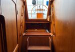 Wooden boat cabin interior with stairs, storage shelves, and a cockpit view showing a steering wheel through an open hatch.