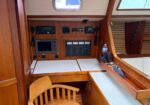 Wood-paneled boat cockpit showing navigation instruments, gauges, and a desk, with a wooden chair and white-cushioned bench.