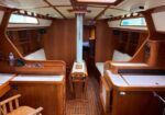 Wood-lined sailboat cabin with white-cushioned benches on both sides and a central mast column.