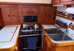 Wood-paneled boat galley with a gas stove, kettle on top, double sink to the right, and shelves of mugs and supplies.