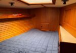 Wood-paneled boat cabin with a blue quilted bed along the left wall and a small hatch at the far end.