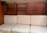Interior of a small boat cabin featuring a white three-seat cushioned bench, wooden cabinets above, and a slatted wooden wall behind.