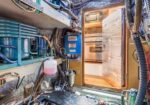 Interior of a small boat engine room with exposed equipment, hoses, and wiring; a door opens to a galley where a fridge and oven are visible.