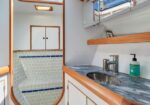 Boat bathroom interior with marble countertop, round sink, soap dispenser, and wooden trim; curved doorway opens to a tiled seating area with a small cabinet above.