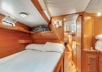 Wooden boat cabin with a single bed on the left, white linens, overhead shelf, small round portlight, and a narrow hallway ahead to other cabins.
