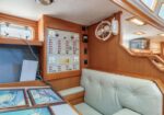 Interior of a boat cabin featuring a wooden control panel with switches and gauges beside a beige cushioned bench seat and a windowed desk area. A white wall fan sits above the panel.