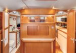 Wood-paneled sailboat cabin with a central divider, navigation desk, and a galley to the right; bathroom visible on the left.