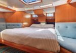 Cozy boat cabin with a large bed, light quilt, wooden walls, blue upholstered benches along the sides, and small porthole windows plus a skylight.