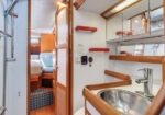 Interior of a small boat cabin featuring a stainless sink with chrome faucet, wooden trim and shelves, mirrored cabinet, and a doorway opening to a sleeping area with a bed and blue cushions.