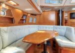 Indoor dining area in a wooden yacht cabin: light-blue cushioned wraparound seating around a polished wooden table on a pedestal, wood panel walls, portholes, and skylights.