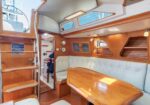 Sailboat cabin with wood paneling, beige seating around a polished wooden table, and stairs to the deck on the left.