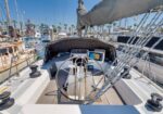 View from the helm of a sailboat docked at a sunny marina; wheel, navigation screen, lines, and other boats in the background.