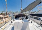 View from the cockpit of a white sailboat at a sunny marina, with other boats and palm trees in the background.