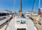 View from the deck of a sailboat in a crowded marina, with tall masts, white hulls, and a clear blue sky now overhead.