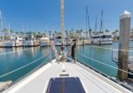View from the bow of a sailboat docked in a marina, with yachts and palm trees along the docks under a clear blue sky.