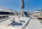 White sailboat deck in a marina, with rigging, coiled rope, and neighboring yachts against a clear blue sky.