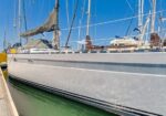 Large sailboat docked at a marina with masts and furled sails under a blue sky; calm green water beside a wooden dock.