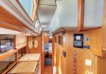Wood-paneled boat interior featuring a compact galley on the left with a stove and microwave, overhead storage, hanging nets, and a cushioned sleeping area toward the back; doorway to a lower cabin visible.