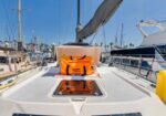 Orange life raft in its open storage hatch on the deck of a white sailboat at a marina, with masts and other boats in the background.