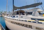 White sailing yacht named Moonraker docked at a marina, with furled mainsail, open cockpit, and blue water in the background.