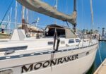 White sailboat Moonraker docked at a marina, with furled mainsail, visible cockpit and rigging against a clear blue sky.