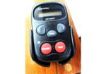 Raymarine S100 remote control with red Standby and Pilot buttons on a wooden surface, featuring gray directional buttons and a small display screen