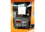 Xantrex Link 20 battery monitor mounted on a wooden panel, showing a red status indicator and a numeric display with buttons labeled Sel, Set, Bat 1, Bat 2.