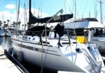 White sailboat named Moonraker moored at a marina, with rigging and other boats in the background and palm trees visible beyond the dock.