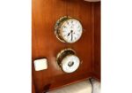 Antique brass clock and barometer mounted on a wooden wall in a boat cabin, with a white light switch nearby.