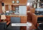 Wood-paneled boat galley with fridge, stove, microwave, and counter, seen from a seating area; stairs lead forward to other sections.