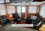 Aft-facing boat salon with wood walls, large windows with blinds, and multiple seating areas including a gray sectional and two recliners, with lamps and pillows for a cozy feel.