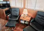 Cozy seating area with two black leather recliners, an ottoman, and a wooden side table with a lamp between them; TV on a cabinet in back.","Two black leather lounge chairs and a matching ottoman sit beside a small wood table holding a lamp and magazines, under window blinds.","A compact living nook featuring leather recliners, an ottoman, and a lamp-lit side table, with a TV cabinet nearby.","Wood-paneled room showing two black leather chairs, an ottoman, and a side table with a lamp and magazines beneath venetian blinds.