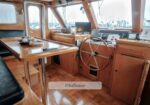 Wood-paneled pilothouse of a boat with a central wheel, navigation instruments and screens, a wooden table with a tea set, and bench seating; marina visible through the windows.