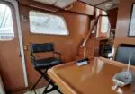 Wood-paneled boat cabin with a rectangular wooden table, a black folding chair, windows letting in light, and stairs leading forward.