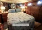 Midship owner’s cabin on a boat with a neatly made bed, blue pillows, and wood paneling; lamps glow beside the bed.