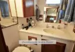 Compact boat ensuite bathroom with a white countertop sink, mirror, and wooden cabinets; towels hang on the wall, toiletries on the counter, and a small window above the vanity.