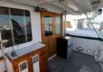 Deck of a yacht with a wooden kitchenette and sink on the left, a small fridge, a wooden door, and a lifebuoy on the railing; marina boats in the background.