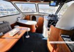 Interior of a boat salon featuring a wooden table, bench seating, and a helm at the front with windows overlooking a marina.