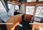 Interior of a boat saloon with helm seat, wooden cabinets and kitchenette, and a seating area; a label notes a full freezer under the helm seat, marina visible through large windows.
