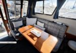 Cozy boat salon with a U-shaped blue-patterned seating around a wooden table, blue curtains, and windows looking out onto a marina.