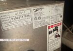 Close-up of a metal water heater with model and serial labels and a red danger/burn warning sticker at the side.