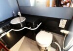 Compact boat bathroom with a white toilet, small sink, black counter, and mirror; reflection of a person taking a photo in the mirror.