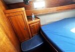 Cozy wooden boat cabin with built-in cabinet, small bedside table, water bottle on top, and a blue-mattress bed beside a cushioned seat.