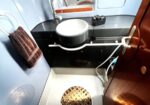 Small boat bathroom: compact washbasin area with circular sink, chrome faucet, mirror, window, towel on a ring, and a floor drain/grate visible near the base of the room.
