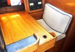 Wooden boat cabin with a navigation desk, blue-cushioned seat, a small desk hole, and a control panel in the background.