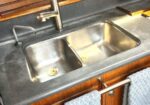 Two blue towels hang from a chrome rack beside a double stainless steel sink in a boat galley, with a dark countertop and wooden cabinets.