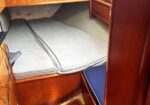 Wood-paneled sailboat cabin with a V-berth mattress and gray cushions, a small wall shelf, and a narrow wooden floor; blue seat to the right.